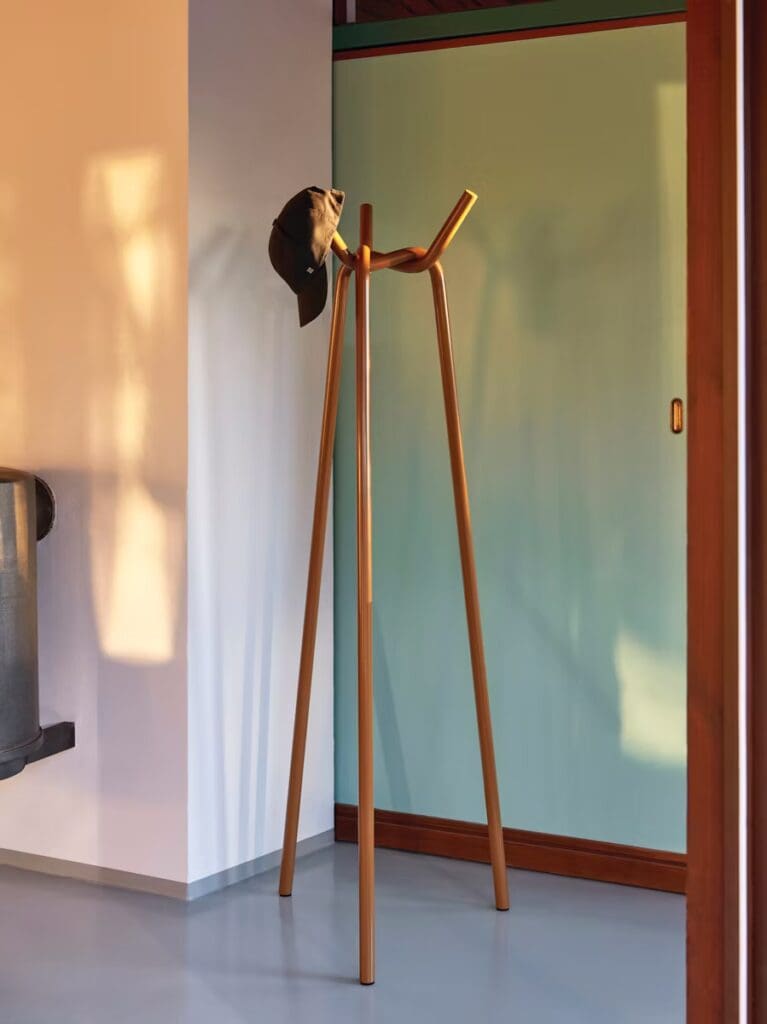 A minimalist coat rack with three upright poles, holding a black hat, situated in a bright room with a green sliding door and a warm light cast on the wall.