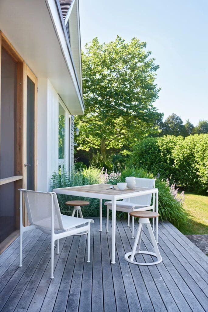 A modern outdoor patio featuring a wooden table with minimalistic design, surrounded by white mesh chairs and stools, set against a backdrop of lush greenery and a bright blue sky.