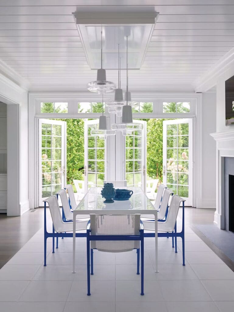 A bright dining area featuring a glass table with blue and white chairs, surrounded by large open windows showcasing greenery outside.