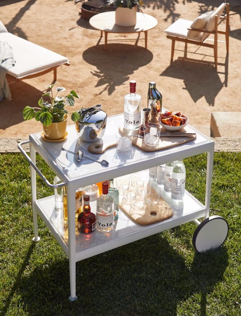 A stylish outdoor bar cart on a grassy area, featuring various bottles of spirits, glasses, a cocktail shaker, and garnishes, with modern lounge furniture in the background.