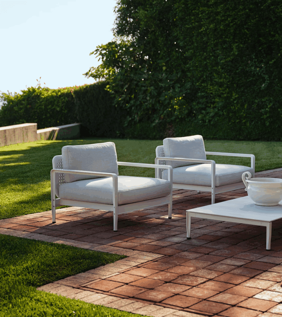 Luxurious outdoor living space featuring two modern white armchairs and a coffee table on a brick patio, surrounded by lush green grass and shrubbery.