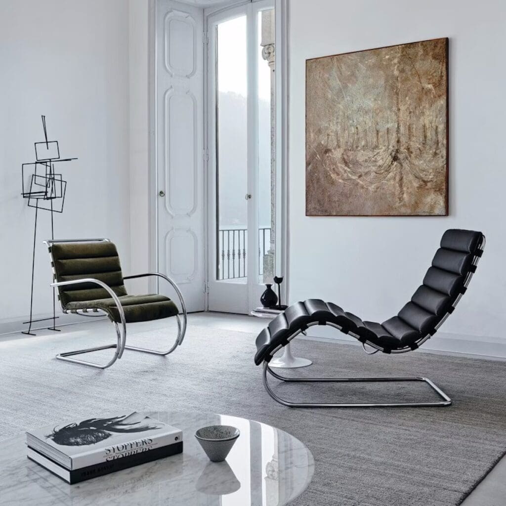 A modern living room showcasing a green chair and a stylish black chaise lounge, both featuring sleek metal frames, with large windows allowing natural light, and an abstract painting on the wall.