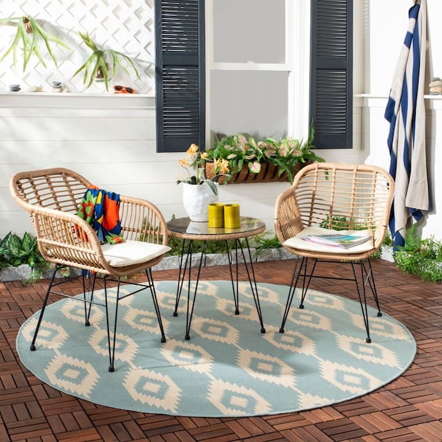 Cozy outdoor seating area featuring two wicker chairs with a round table in between, set on a patterned rug. Lush greenery and potted plants are visible in the background.