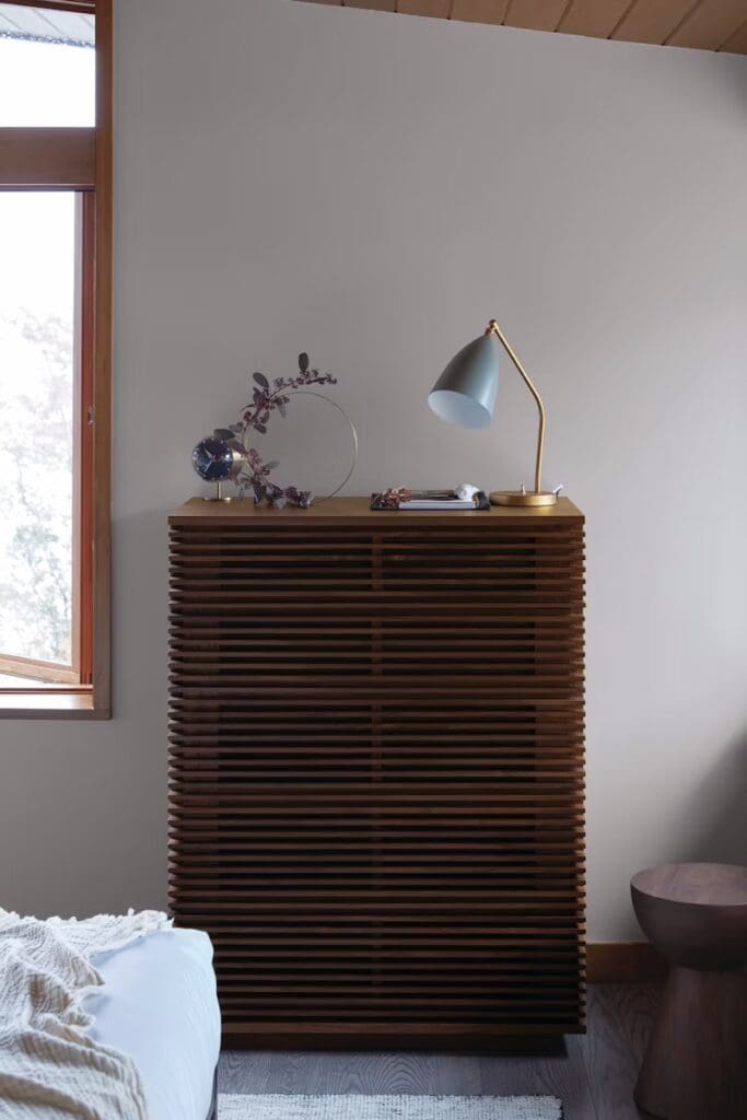 A modern bedroom corner featuring a stylish wooden dresser with horizontal slats, a mint green lamp, and decorative elements, alongside a window allowing natural light.