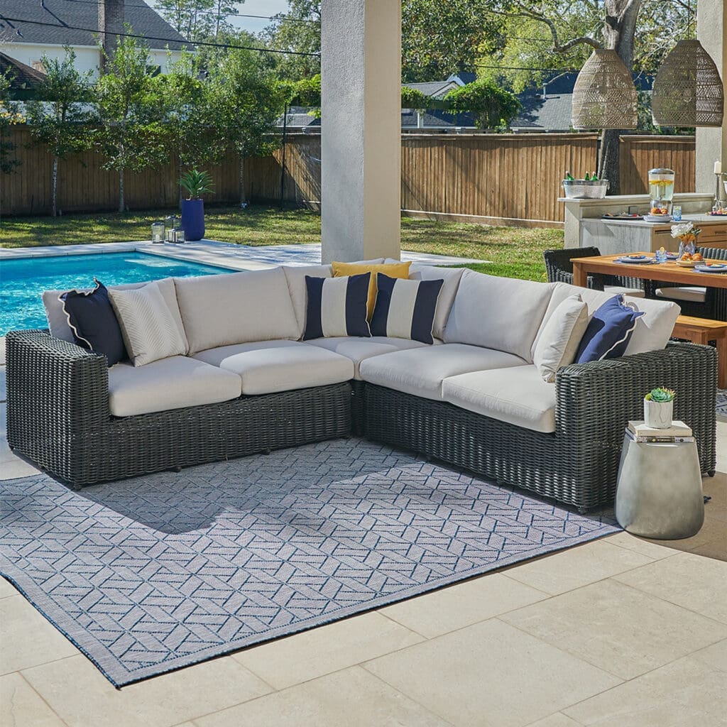 Luxurious outdoor seating area featuring a stylish sectional sofa with navy and white pillows, a decorative rug, and a nearby pool in a sunny backyard.