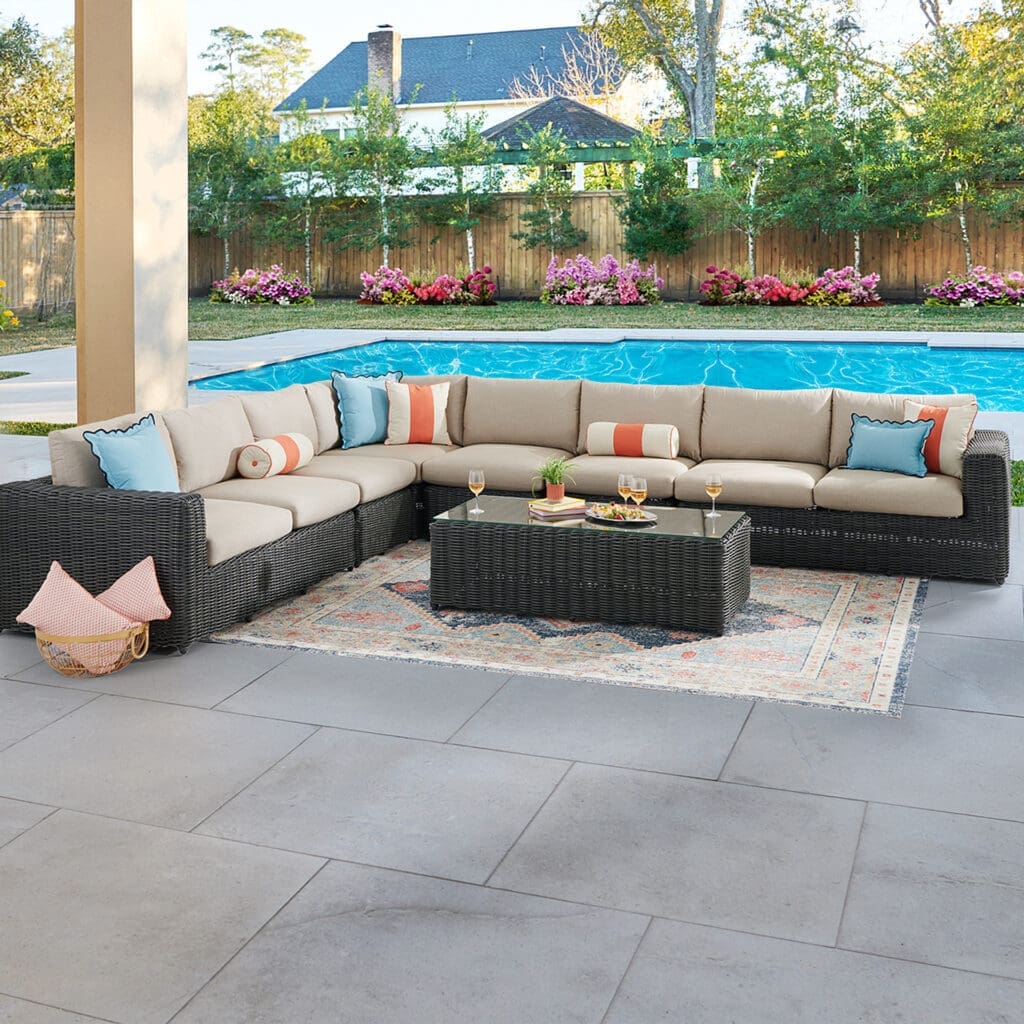 Stylish outdoor living area featuring a large sectional sofa with cushions, a coffee table, and a vibrant area rug, adjacent to a swimming pool and surrounded by lush greenery and flowers.