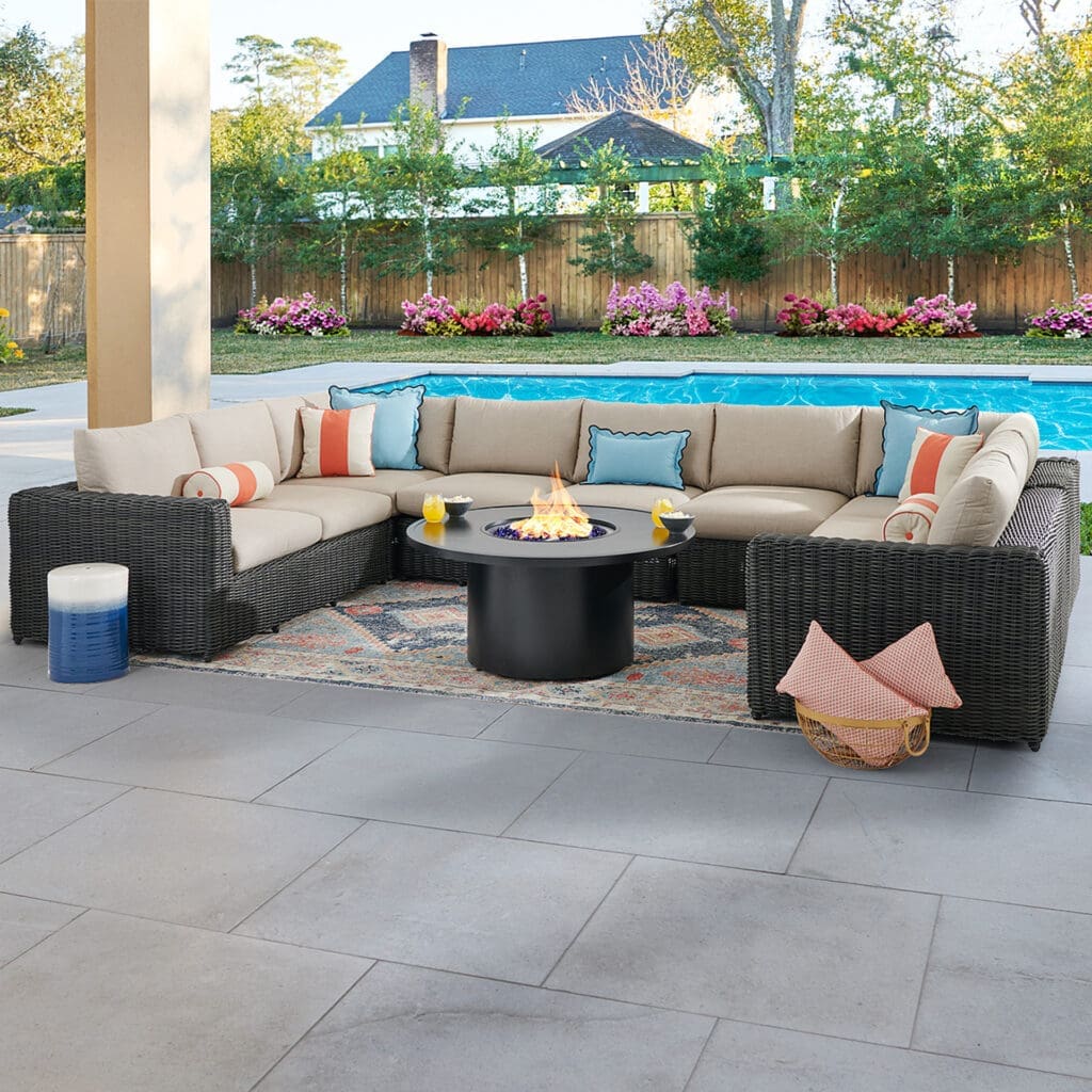 Luxurious outdoor living space featuring a large, curved sofa with light-colored cushions, a central fire pit table, and refreshing drinks on the table, with a swimming pool and flowering plants in the background.
