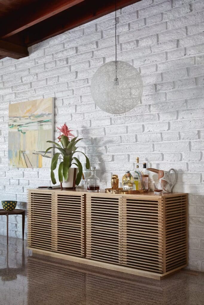 A modern interior featuring a wooden sideboard with slatted design, a vibrant plant, various liquor bottles, and a round decorative light fixture. The wall is made of white bricks, enhancing the contemporary aesthetic.