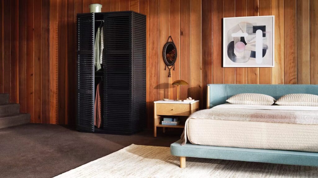 A cozy bedroom featuring a light blue upholstered bed, a wooden nightstand with a lamp, and a modern black wardrobe against warm wooden wall paneling.
