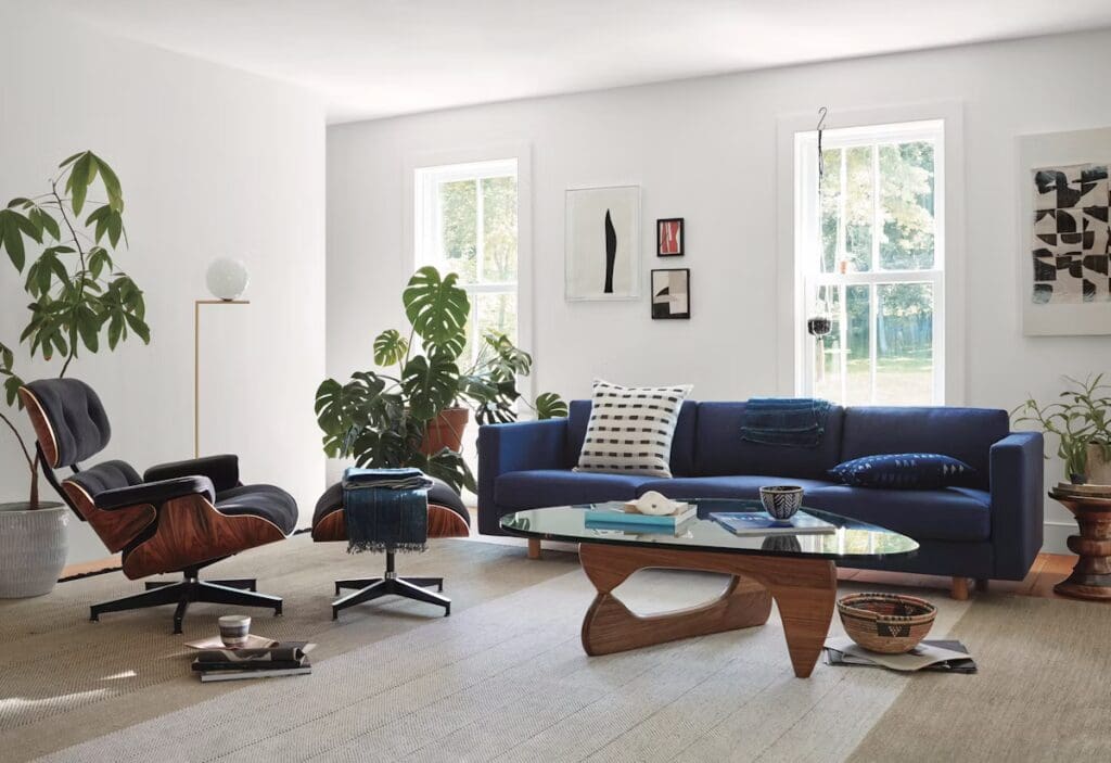 A modern living room with a blue sofa, a stylish wooden coffee table, an elegant armchair, and indoor plants, featuring large windows that let in natural light.