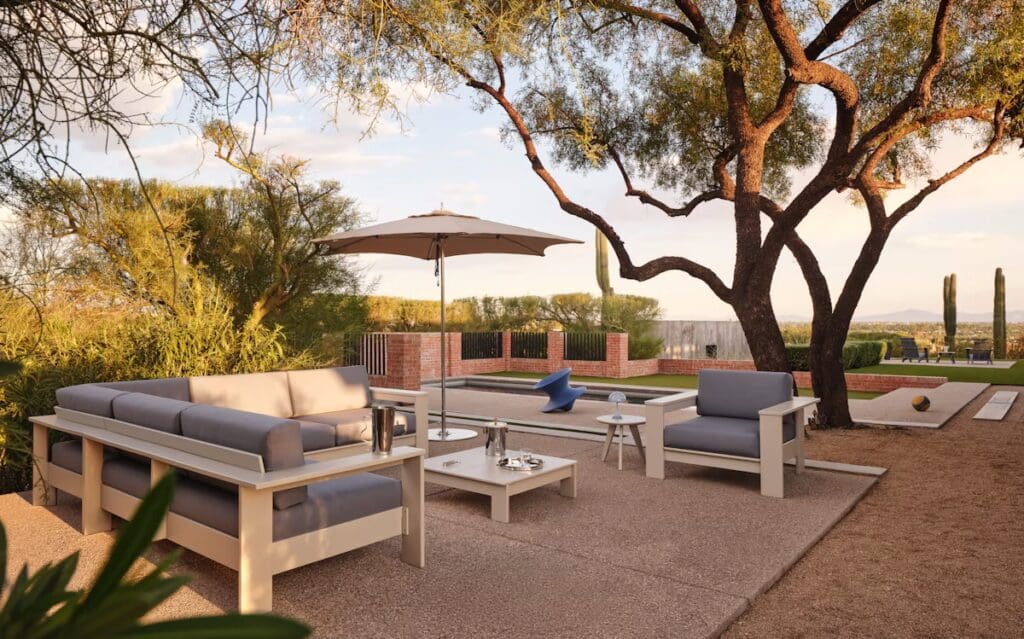A modern outdoor living space featuring sleek gray sofas with cushions, a round table and an umbrella for shade, surrounded by greenery and a scenic view.