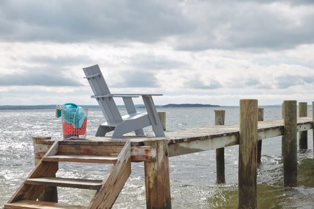 A wooden dock extending over water, featuring a gray Adirondack chair and a wire basket with towels, under a cloudy sky.