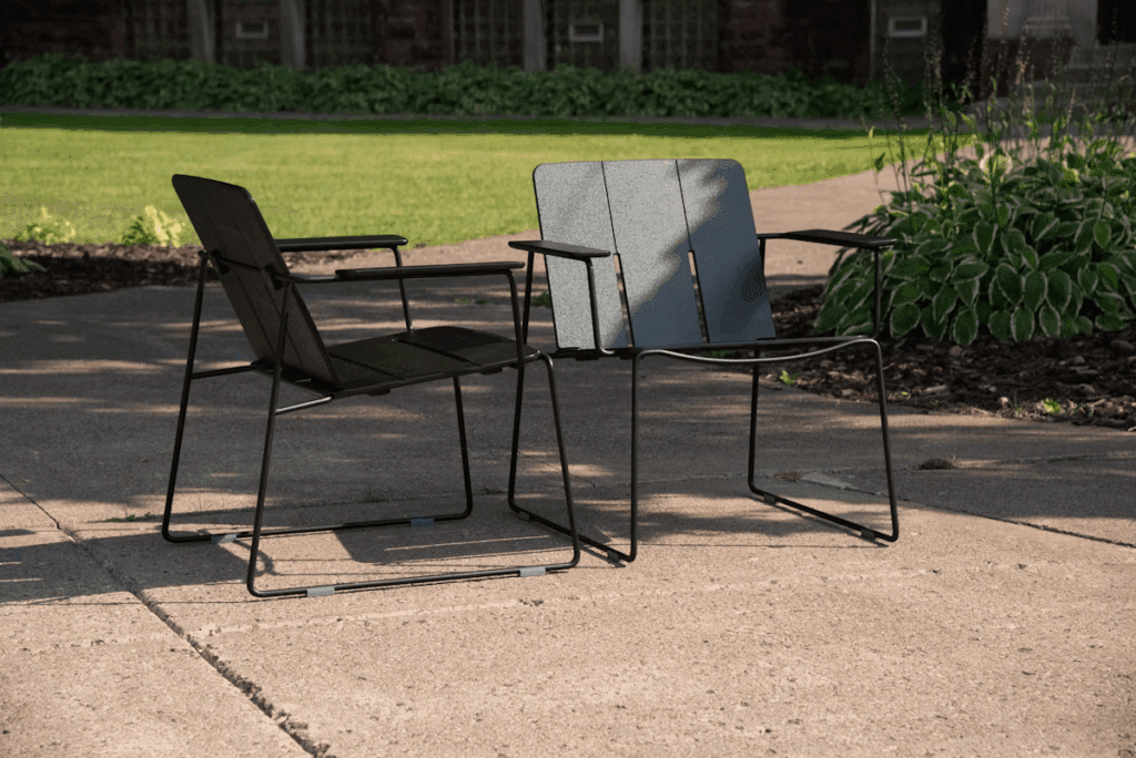Two modern outdoor chairs with a sleek design positioned on a paved surface, surrounded by greenery.