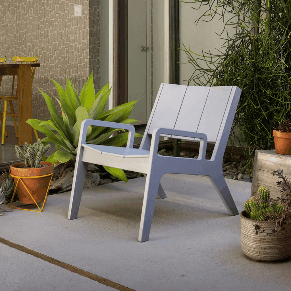 Stylish outdoor patio scene featuring a modern light gray chair next to potted plants, with a wooden table and green foliage in the background.