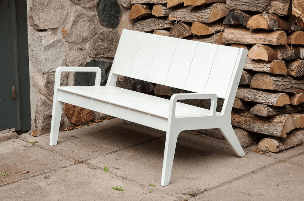 Modern white outdoor bench positioned next to a stone wall and a stack of firewood.