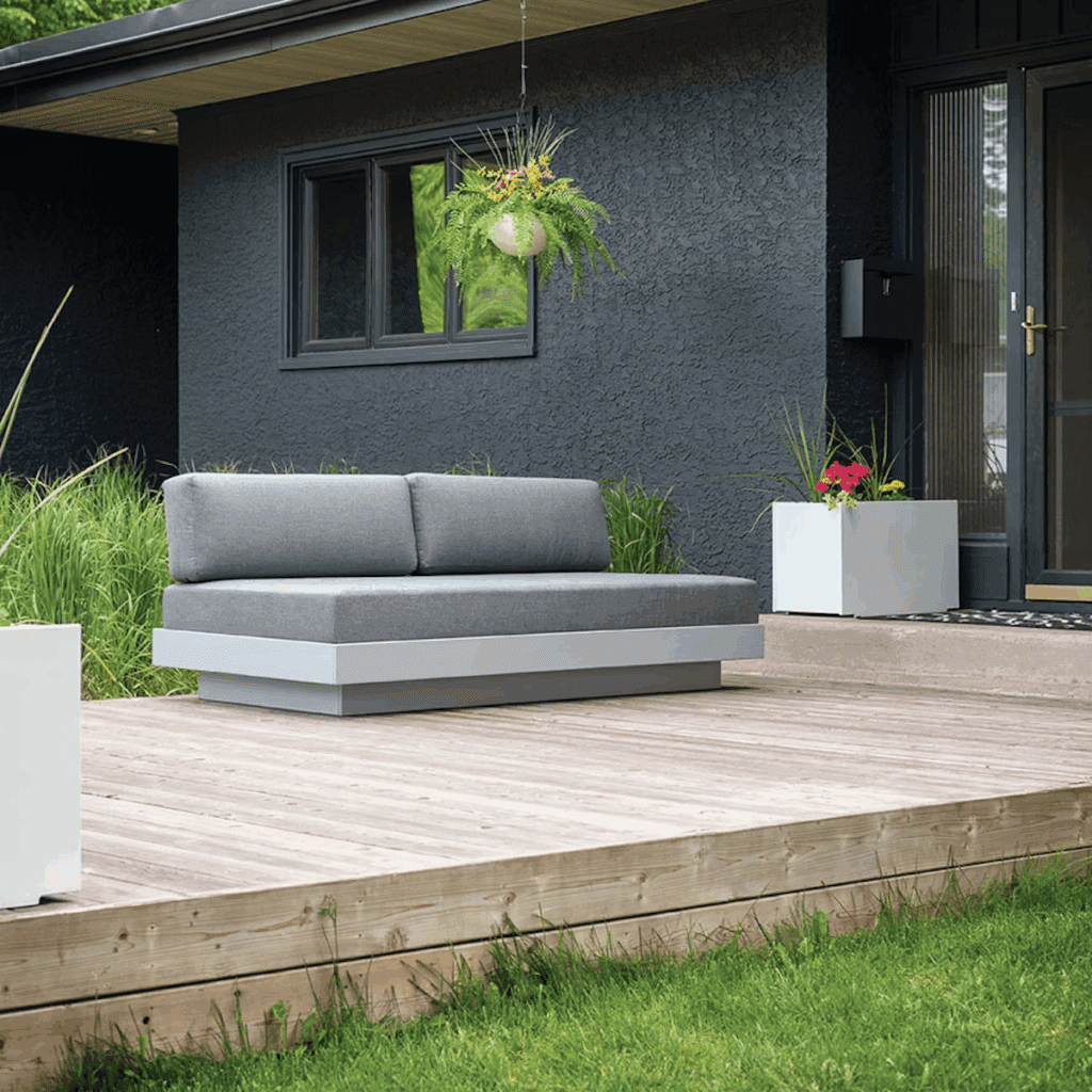 Modern outdoor seating area featuring a grey sofa on a wooden deck, surrounded by greenery and potted plants.