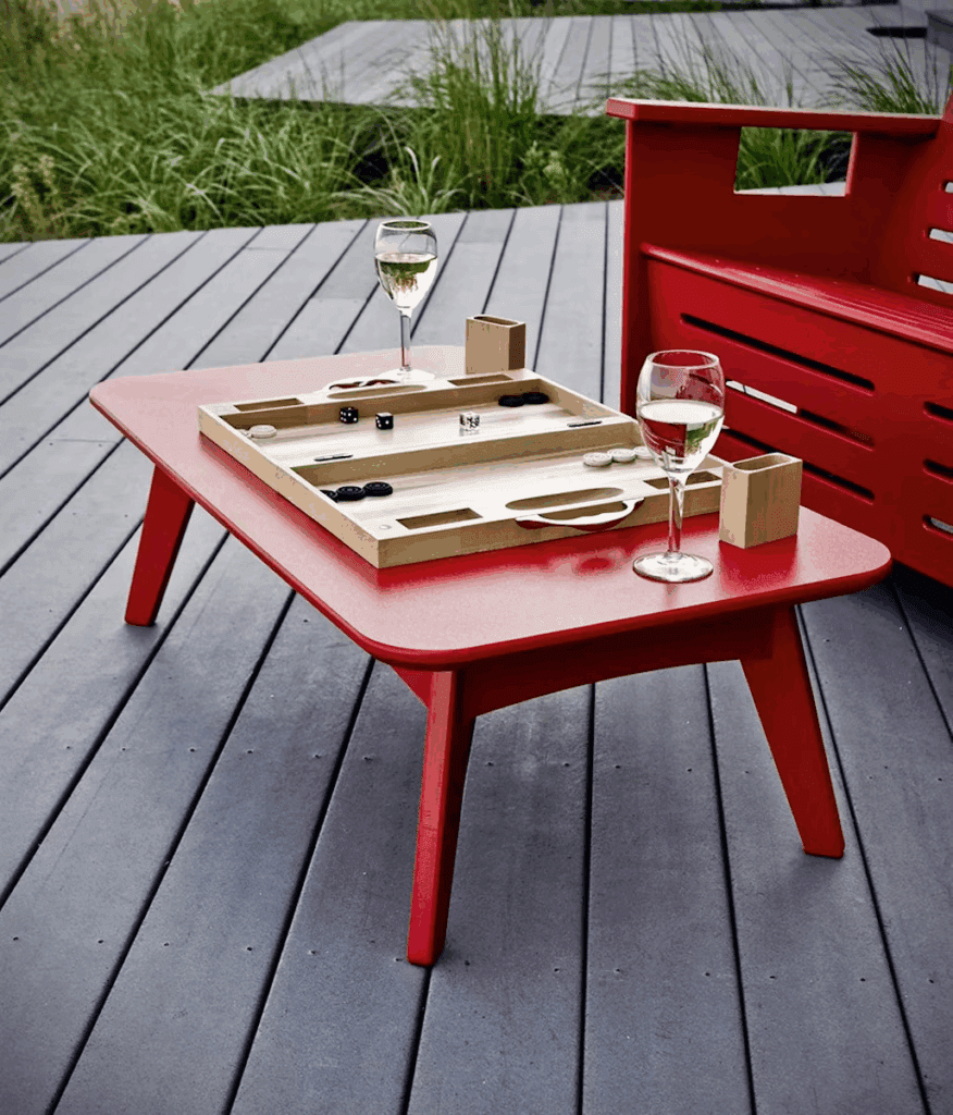 Bright red outdoor coffee table with a backgammon game set, two glasses of wine, and bamboo game pieces, surrounded by lush greenery and a wooden deck.
