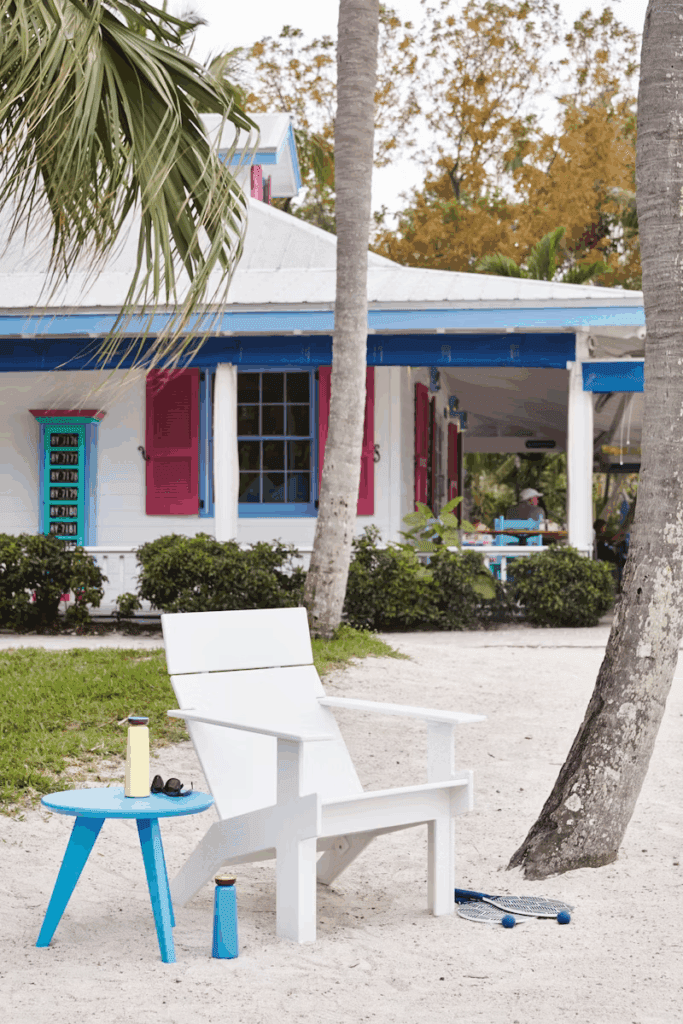 Elegant outdoor seating area featuring a white Adirondack chair and a blue side table, with palm trees and a colorful beach house in the background.