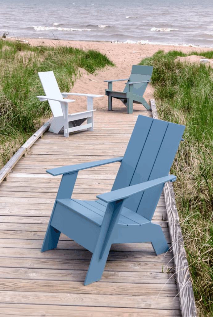 Adirondack chairs in blue and white arranged on a wooden boardwalk leading to a sandy beach, surrounded by grass and a calm ocean.