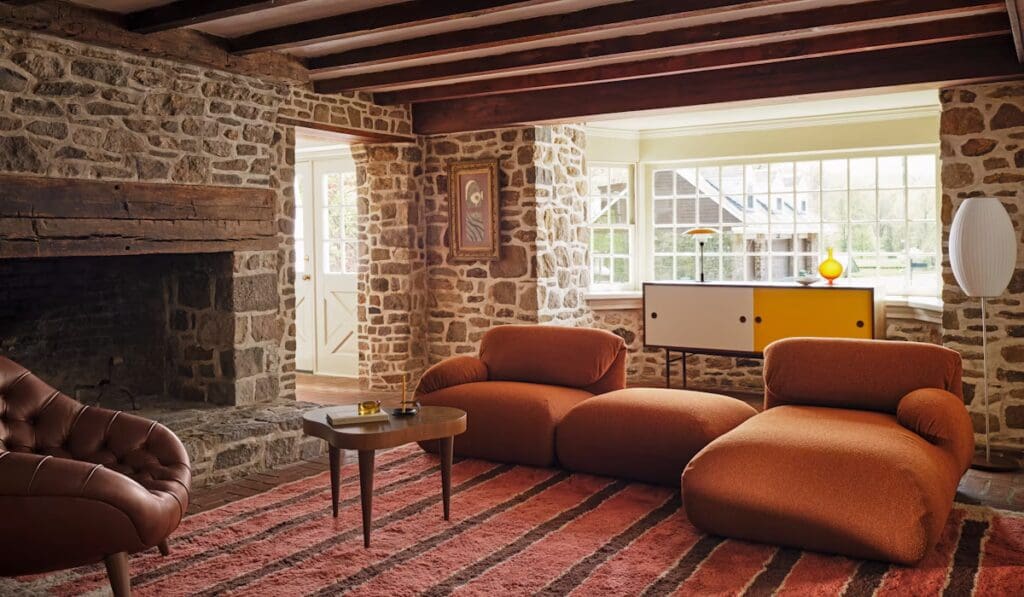 A cozy living room featuring a stone wall, a modern orange sofa, a round coffee table, and a stylish brown armchair. Natural light streams through large windows, illuminating the space.