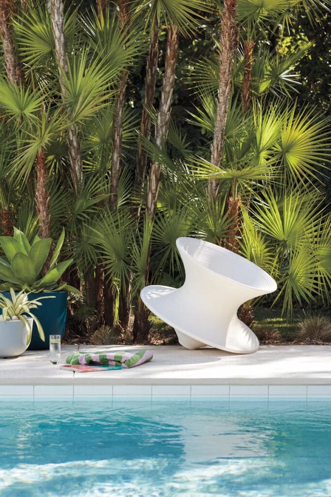 A modern white spiraled chair positioned beside a swimming pool, surrounded by lush green plants and a colorful towel next to a glass of water.
