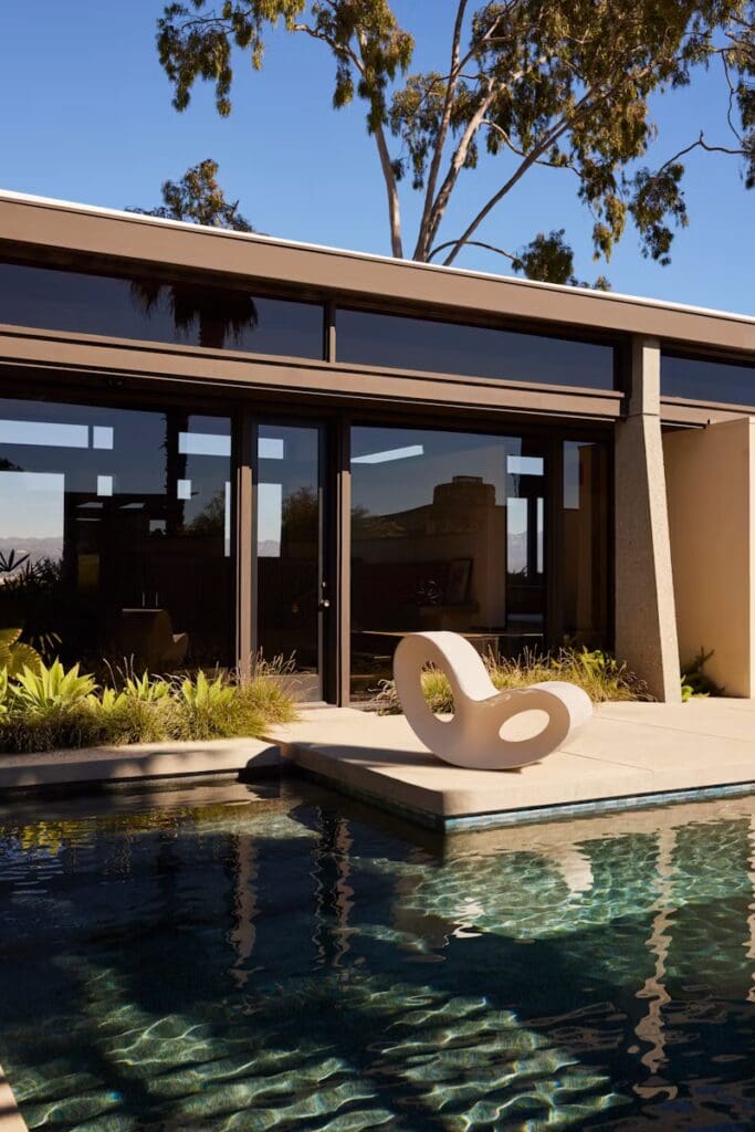 A modern outdoor scene featuring a unique abstract chair next to a swimming pool, surrounded by lush greenery and a contemporary house with large windows.