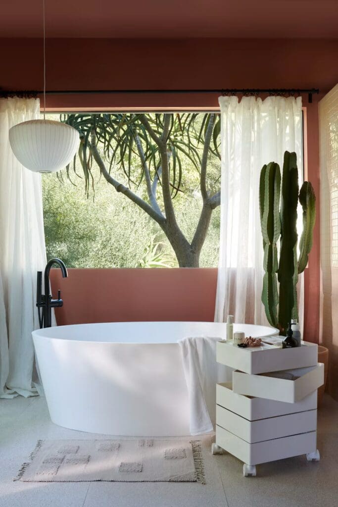 A stylish bathroom featuring a white freestanding bathtub, a modern black faucet, and a small white storage unit on wheels. Natural light pours in through a large window adorned with sheer curtains, revealing a lush view of greenery outside.