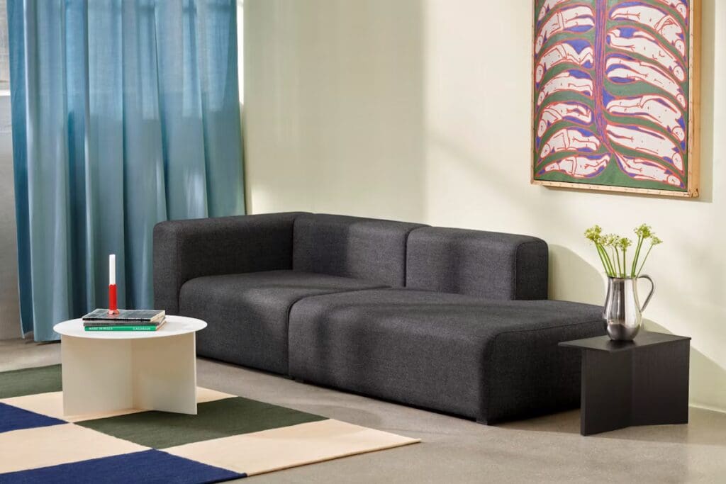 A modern living room with a dark gray sectional sofa, a minimalist white coffee table, and a small black side table. The decor includes a colorful artwork on the wall and green curtains draping over a window.