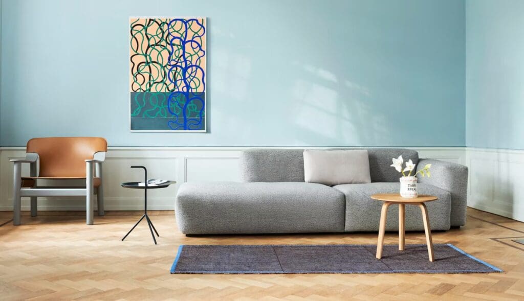 A modern living room featuring a gray sofa with a single cushion, a small round side table with a vase of flowers, and a stylish armchair. The wall is painted light blue and decorated with abstract art.