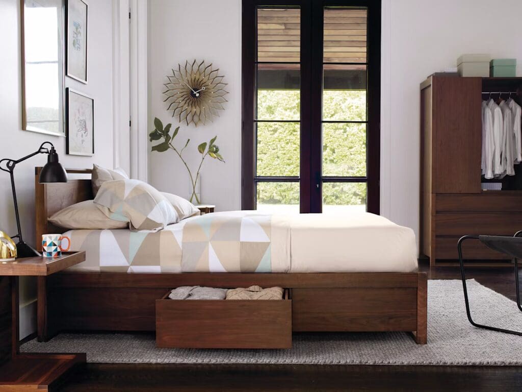 A modern bedroom featuring a wooden bed with a geometric-patterned duvet, a bedside table with a lamp and decorative mug, and large windows providing natural light. The room includes a wardrobe and a rug, creating a cozy and stylish atmosphere.