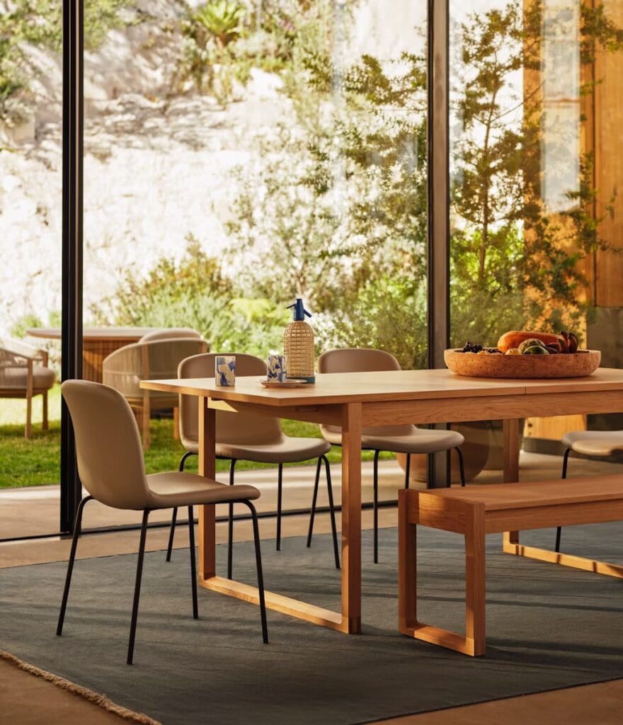 A modern dining area featuring a wooden table, coordinating chairs, a decorative fruit bowl, and large windows overlooking a green garden.