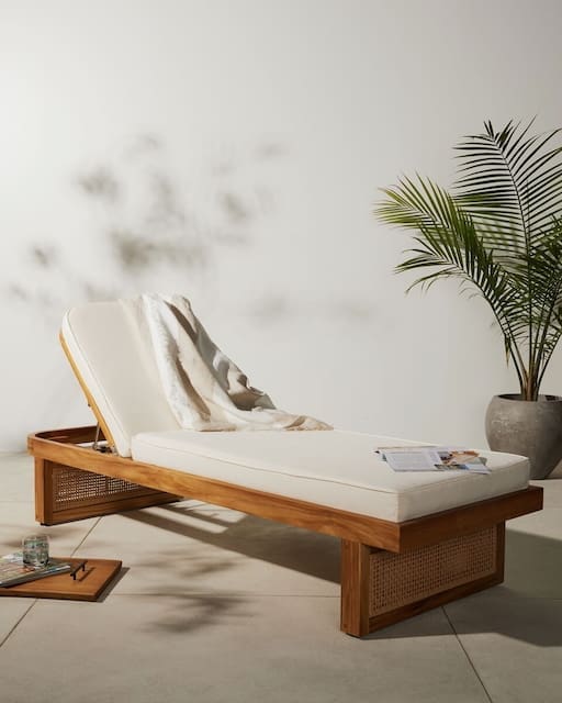 A modern wooden lounge chair with a white cushion and a cozy blanket, positioned next to a decorative palm plant, against a neutral wall.
