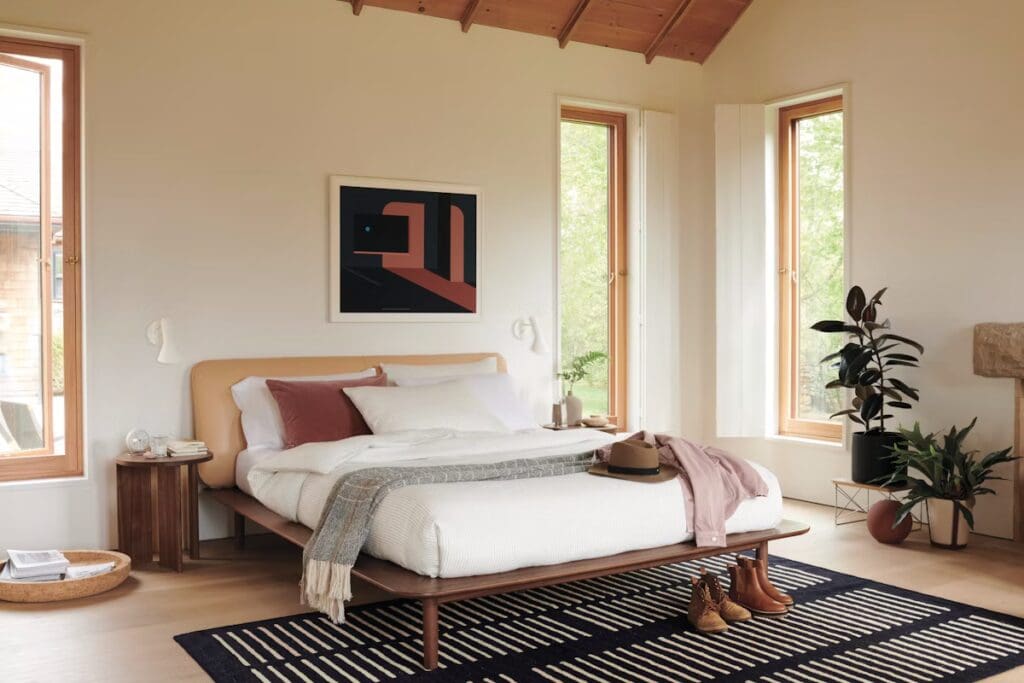 A modern bedroom featuring a bed with a light-colored upholstered headboard, decorative pillows, and a cozy throw blanket. The room has large windows with wooden frames allowing natural light, a bedside table with a decorative lamp, and a stylish rug on the floor. Plants are placed in the corners, creating a fresh and inviting atmosphere.