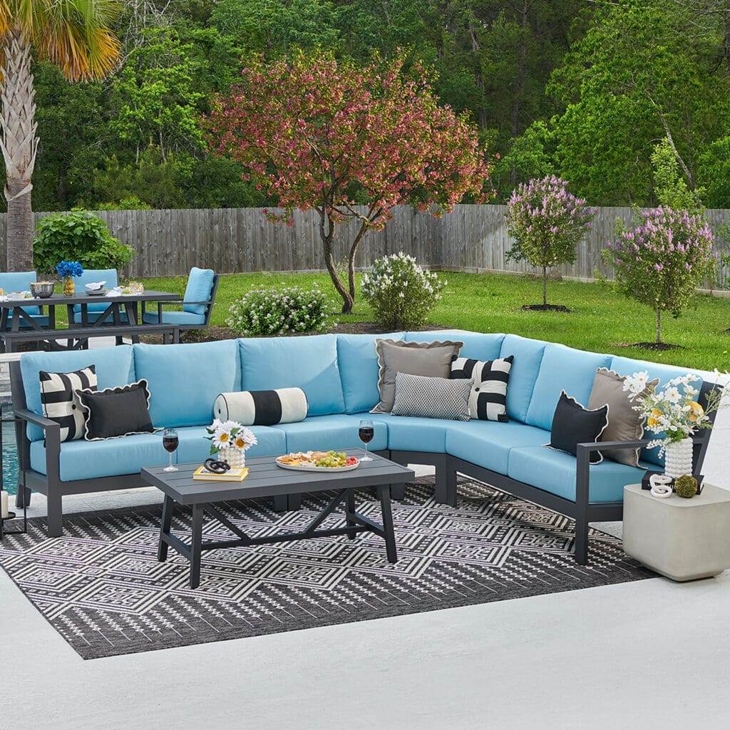 Stylish outdoor seating area featuring a blue sectional sofa with patterned cushions, a black coffee table, and decorated with flowers and snacks, set in a green backyard.