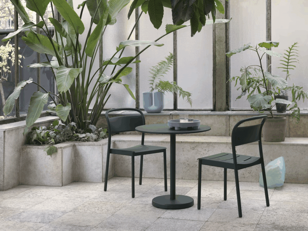 Elegant outdoor dining area featuring a round table and two chairs surrounded by lush tropical plants, set in a bright and airy space.