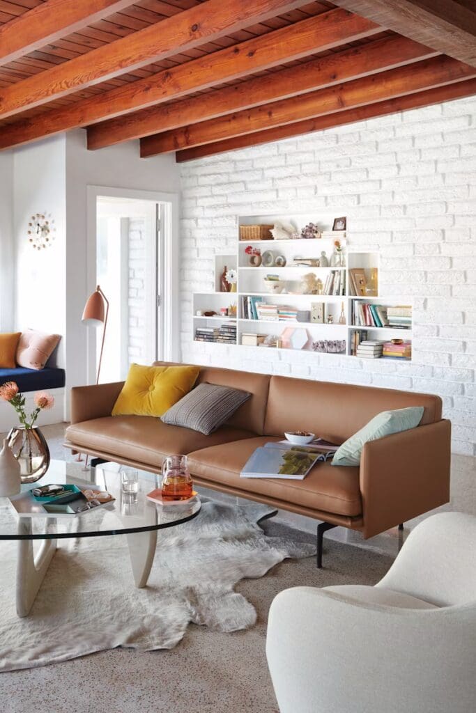 A modern living room with a brown sofa adorned with colorful cushions, a round glass coffee table on a light rug, and a stylish floor lamp. The walls are white brick, and there's a shelving unit displaying books and decor items. Natural light floods through large windows.