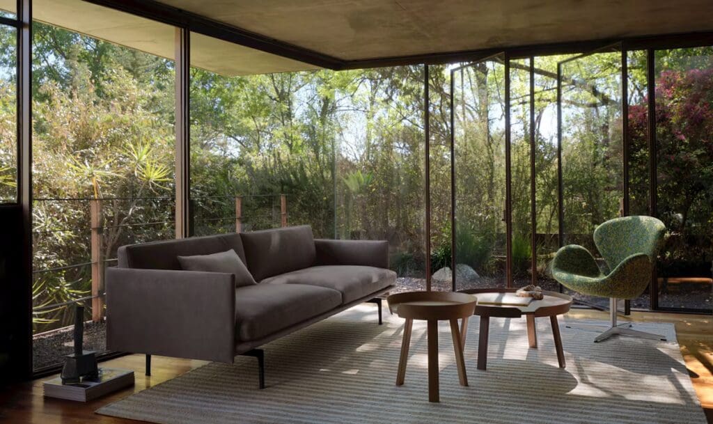A modern living room featuring a gray sofa, circular wooden coffee tables, and a patterned accent chair, surrounded by large glass windows showcasing greenery outside.