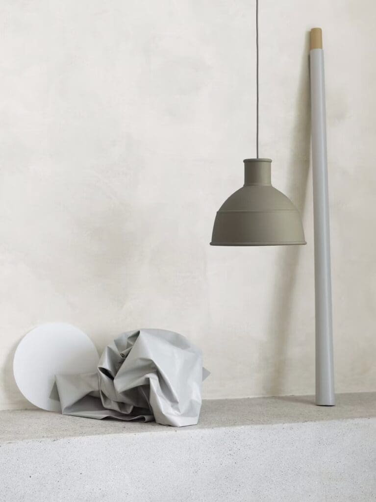 A minimalist interior scene featuring a grey pendant light hanging above a textured, light-colored surface with a crumpled grey fabric and a round white object beside a tall, slender white pole.