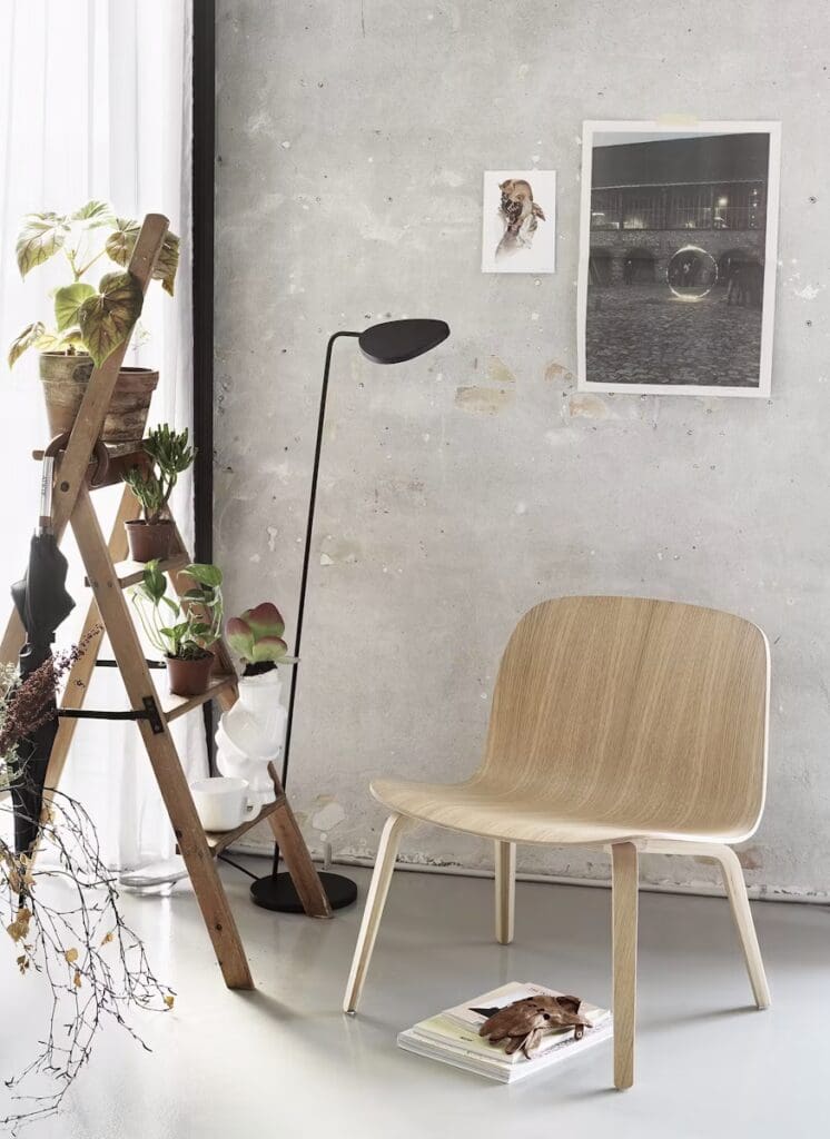 A minimalist living space featuring an elegant wooden chair, a floor lamp, and a plant display on a ladder shelf, with grey concrete walls and artwork on display.