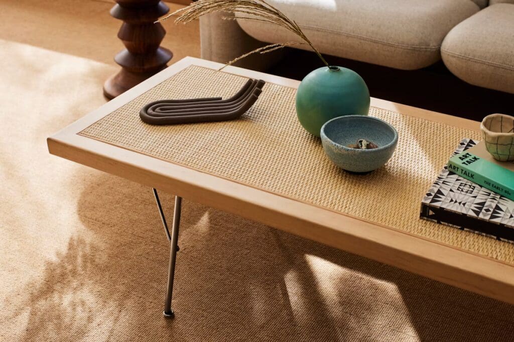 A close-up view of a wooden coffee table with a natural woven surface, featuring a decorative brown coiled object, a green decorative ball, a small blue bowl, and a book titled 'ART TALK' surrounded by soft natural lighting.