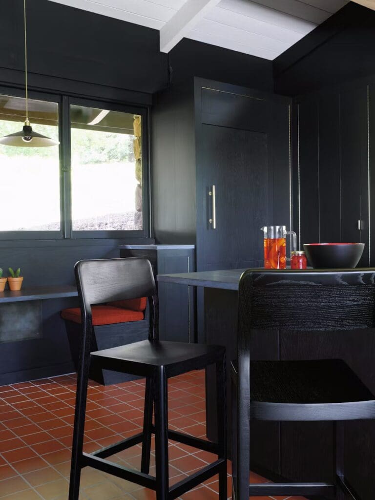A modern kitchen featuring black walls and cabinetry, a wooden table with black stools, and a large window providing natural light. Decorative elements include a pendant light and potted plants on a ledge.