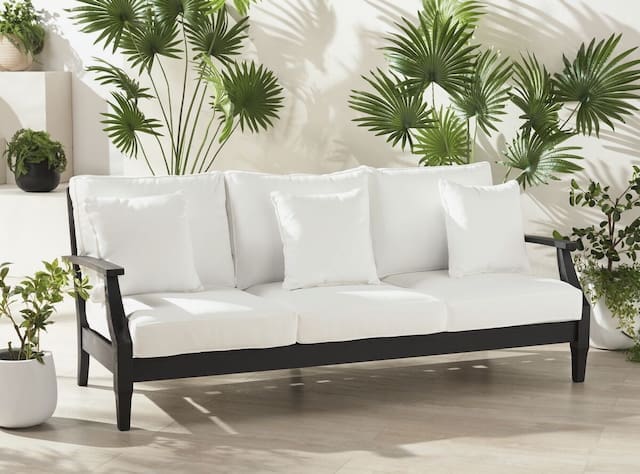 Modern outdoor sofa with white cushions, surrounded by lush green plants in a bright setting.