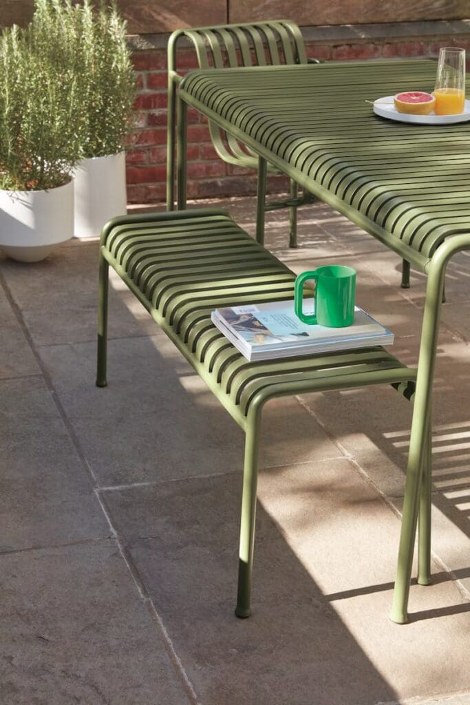 A green outdoor dining set consisting of a table and a bench with slatted design, accompanied by a green mug and a magazine on the bench. In the background, potted greenery is visible against a brick wall.