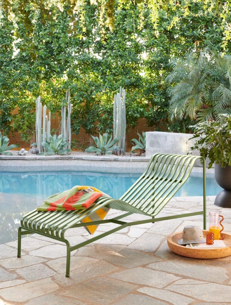 A green lounge chair with a colorful towel draped over it, placed beside a swimming pool surrounded by lush greenery. A sandy-colored round table holds a hat and a drink.