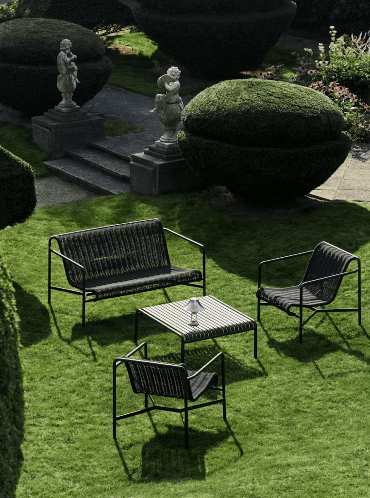Elegant garden seating area featuring black metal chairs and table on lush green grass, surrounded by neatly trimmed hedges and decorative statues.