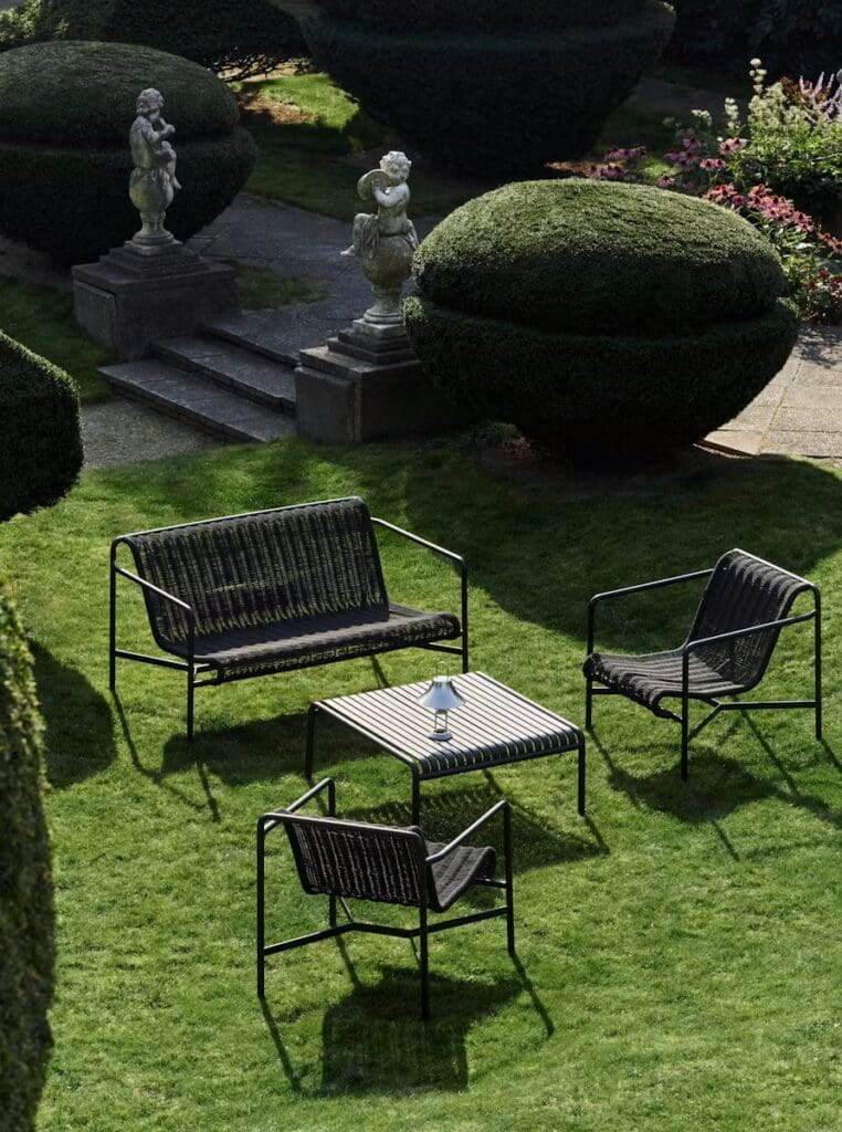 A serene garden scene featuring a black metal outdoor seating set, consisting of two chairs and a long bench around a striped coffee table, surrounded by lush green grass and neatly trimmed hedges, with classical sculptures in the background.