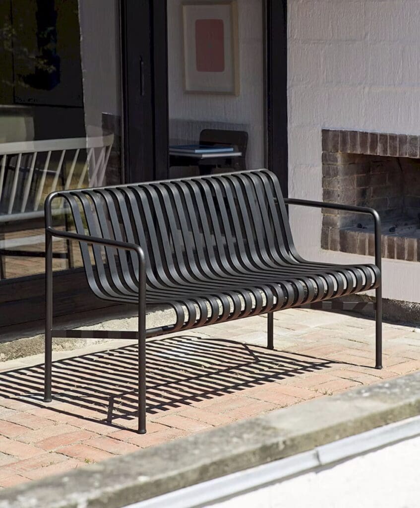 A modern outdoor bench with horizontal slats and a sleek metal frame, situated on a brick patio with a view of large windows and a fireplace.