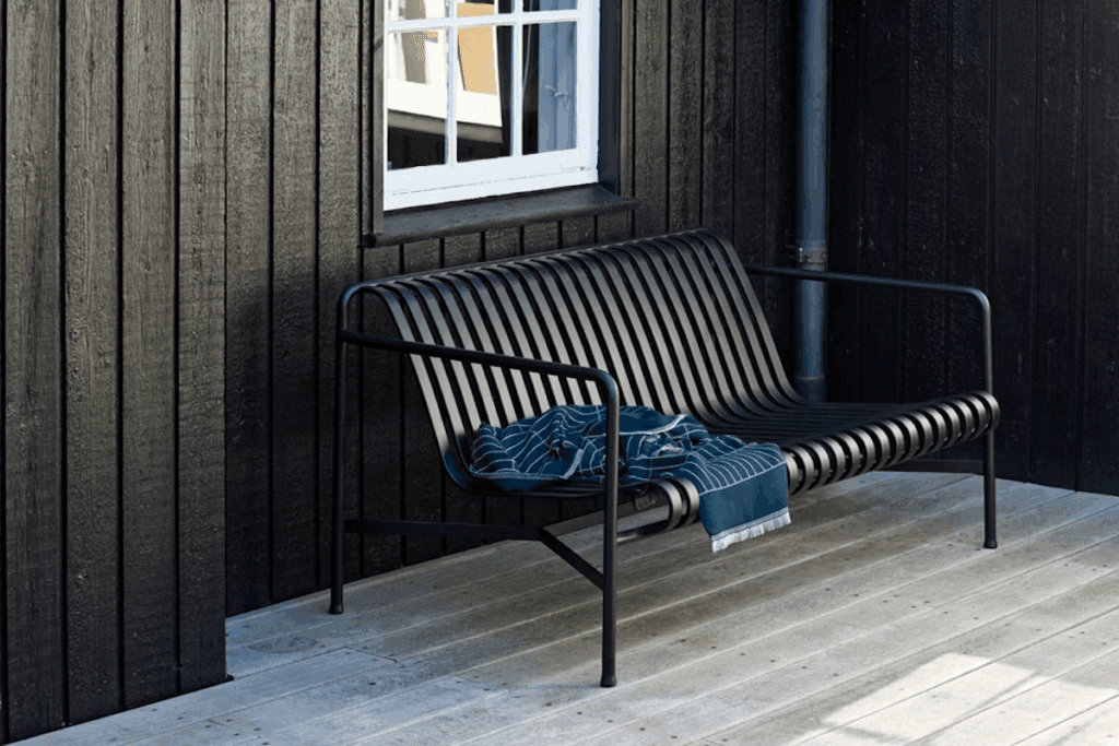 Modern black outdoor bench with a striped design, adorned with a blue throw blanket, set against a dark wooden wall.