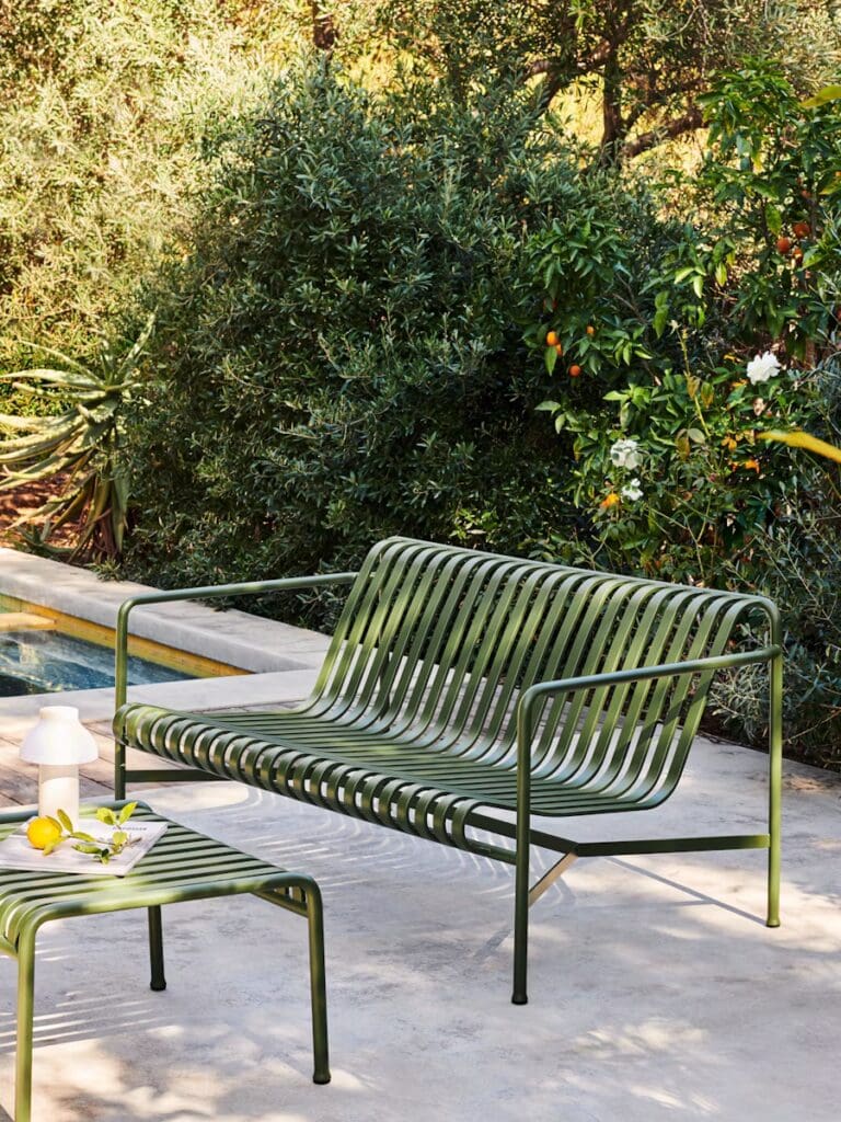 A green metal sofa and matching side table are positioned near a swimming pool, surrounded by lush greenery.
