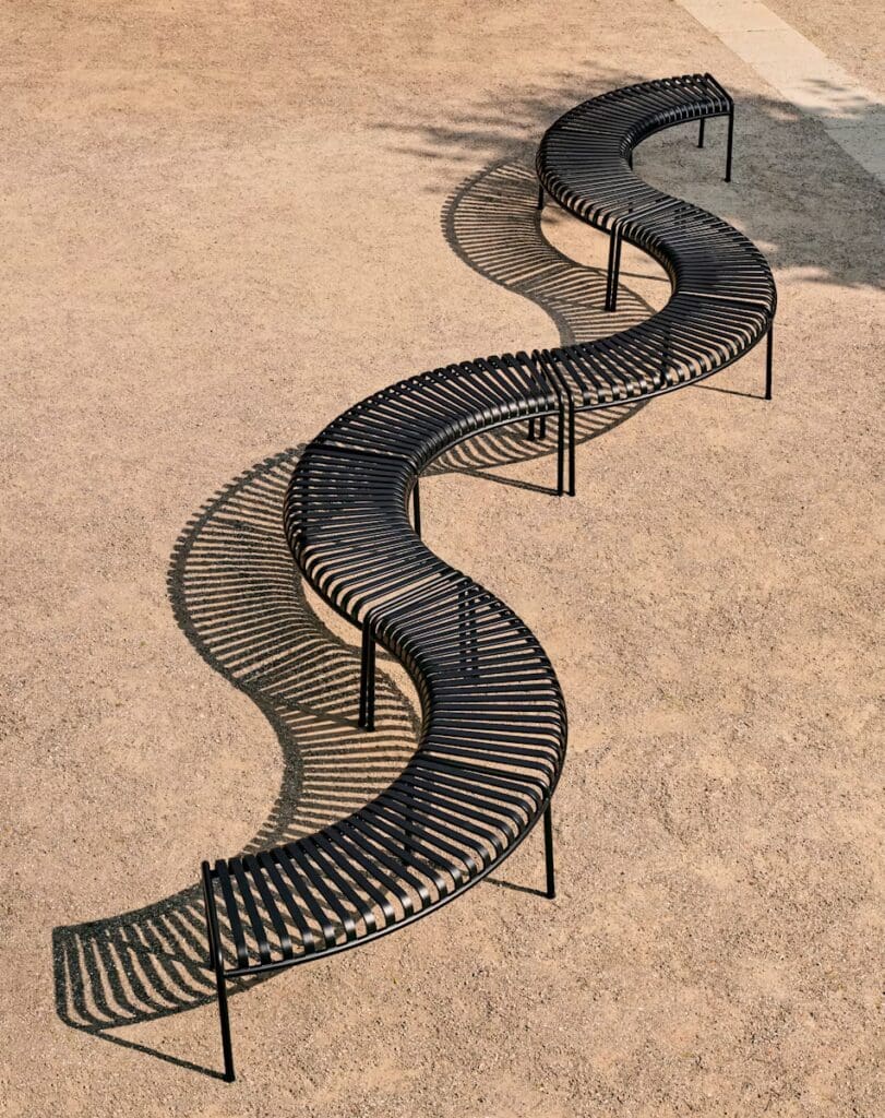 A sleek, wave-shaped black bench made from slatted material, set against a sandy ground, casting intricate shadows.